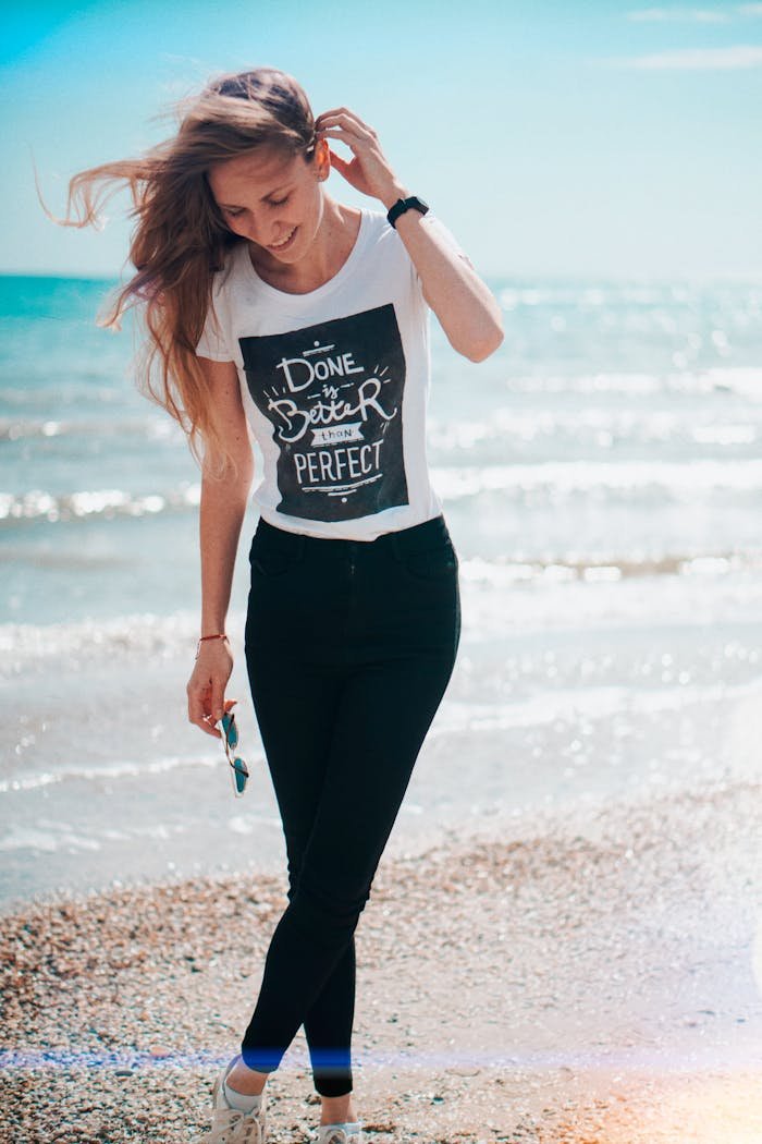 A woman walks on a sunlit beach in casual clothing, embodying a carefree summer vibe.